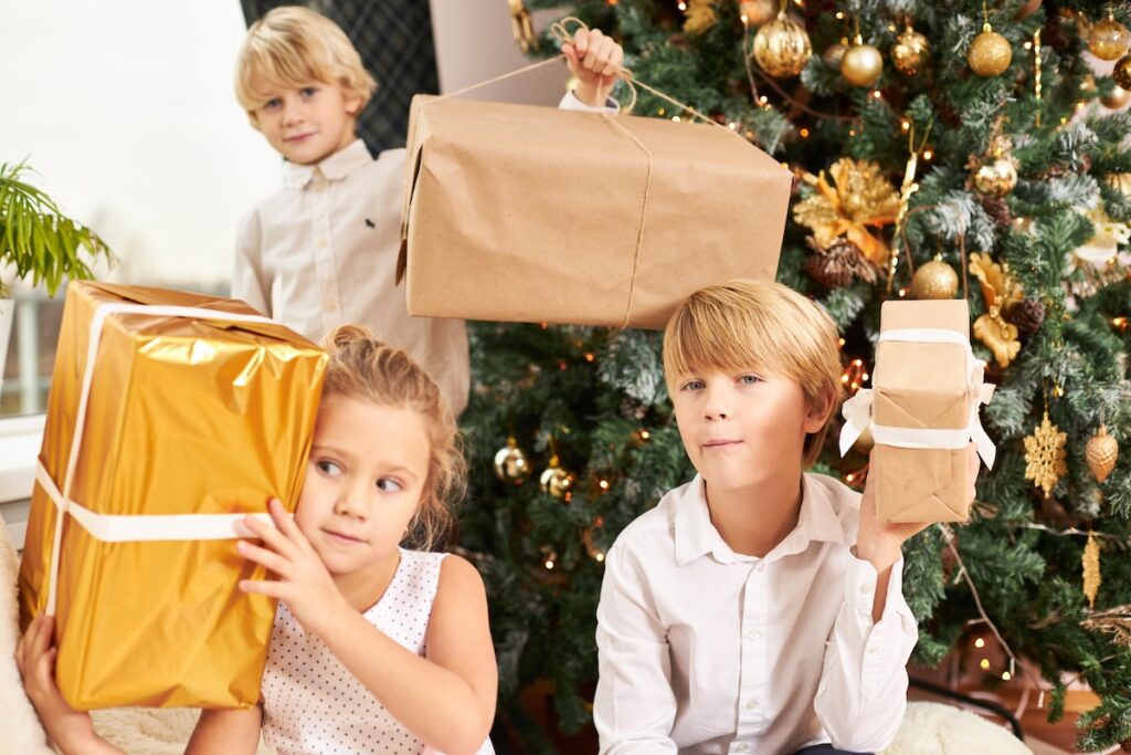 horizontal shot three cute siblings sitting decorated new yeara s tree holding boxes with christmas presents feeling impatient having curious looks happy childhood joy festivity