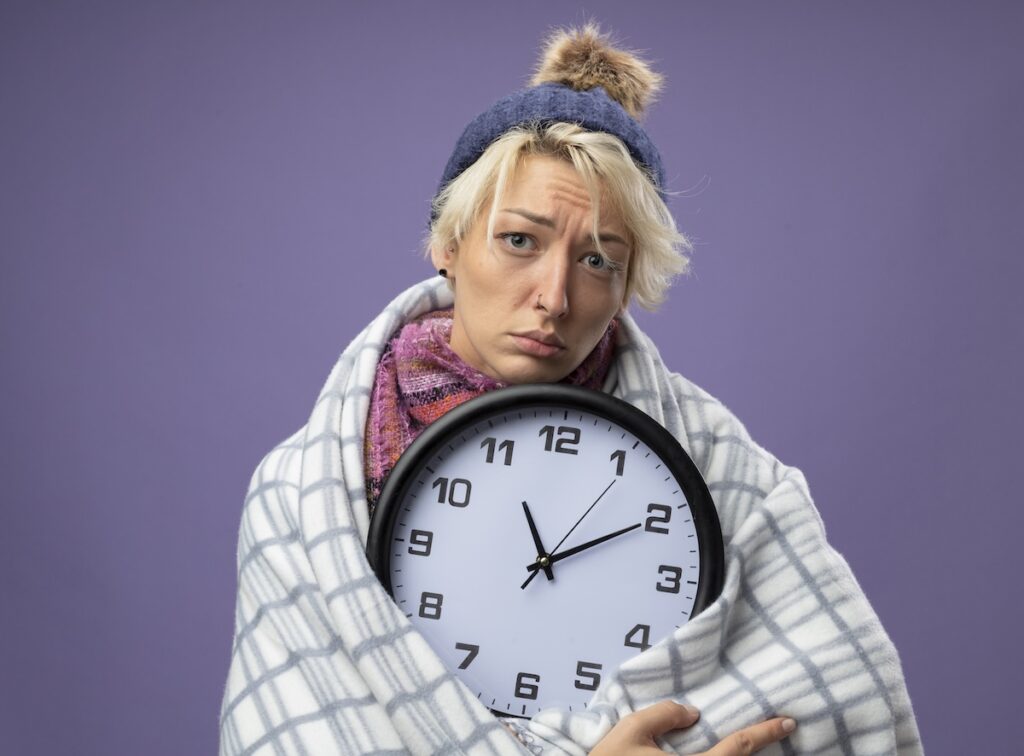 sick unhealthy woman with short hair warm scarf hat feeling unwell wrapped blanket holding wall clock looking camera with sad expressionover purple background