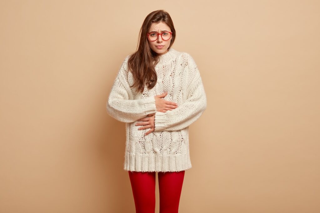 upset sullen european female touches belly from pain feels unwell discomfort after eating spoiled product wears spectacles warm clothes stands brown wall stomachache concept