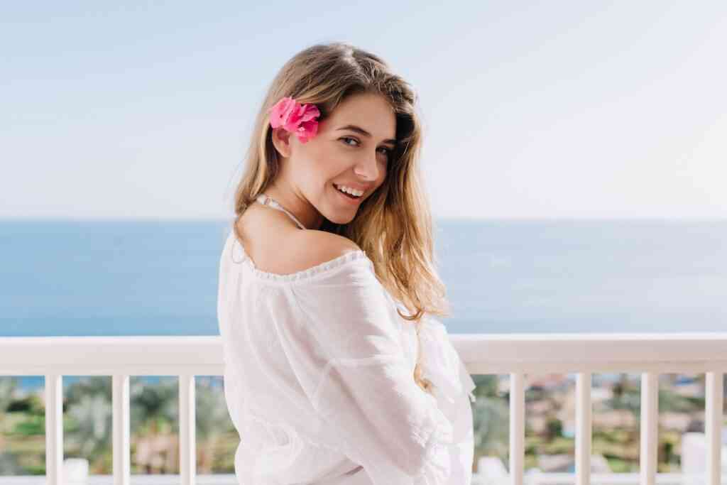 cute happy girl with pink flower her hair smiling enjoys summer vacation resort portrait cheerful young woman white shirt standing terrace amazing ocean views